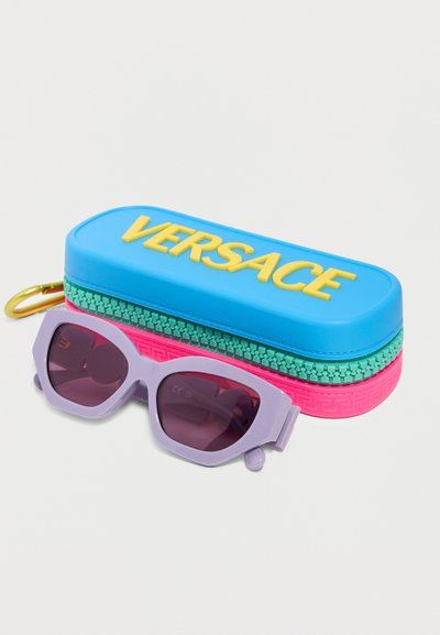 Μωβ γυαλιά ηλίου με φακούς σε σχήμα γατίσιου ματιού δίπλα σε πολύχρωμη θήκη Versace σε μπλε, πράσινο και ροζ με κίτρινα γράμματα.