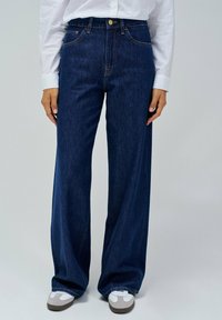 Jeans in denim blu scuro a gamba larga, con vita alta, dotati di tasche frontali e chiusura a bottoni. Abbinate a sneakers grigie e bianche.