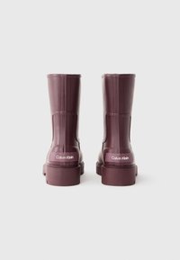 Bottes en caoutchouc bourgogne Calvin Klein avec une semelle plate, texture lisse et logo au dos. Hauteur mi-mollet avec un design à coupe droite.