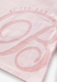 Růžové bavlněné tričko s velkým, světle růžovým stylizovaným grafickým prvkem "R" a decentním značkovým textem nahoře. Hladká textura, krátké rukávy.