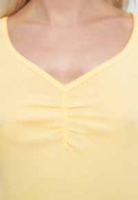 Żółty, prążkowany top z dekoltem w serek, z marszczeniem na biuście i falistymi brzegami przy szyi. Miękka faktura, dopasowany krój.
