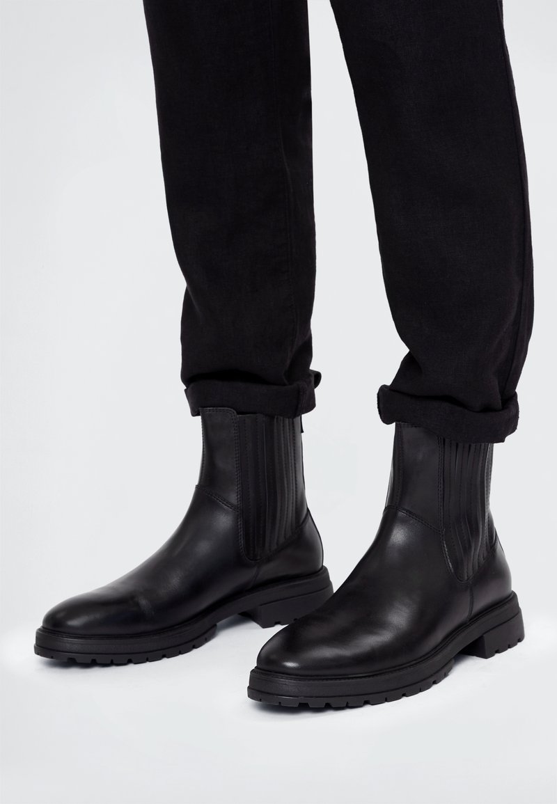 Stivaletti neri in pelle con pannelli elastici laterali indossati con pantaloni neri arrotolati su uno sfondo bianco semplice.