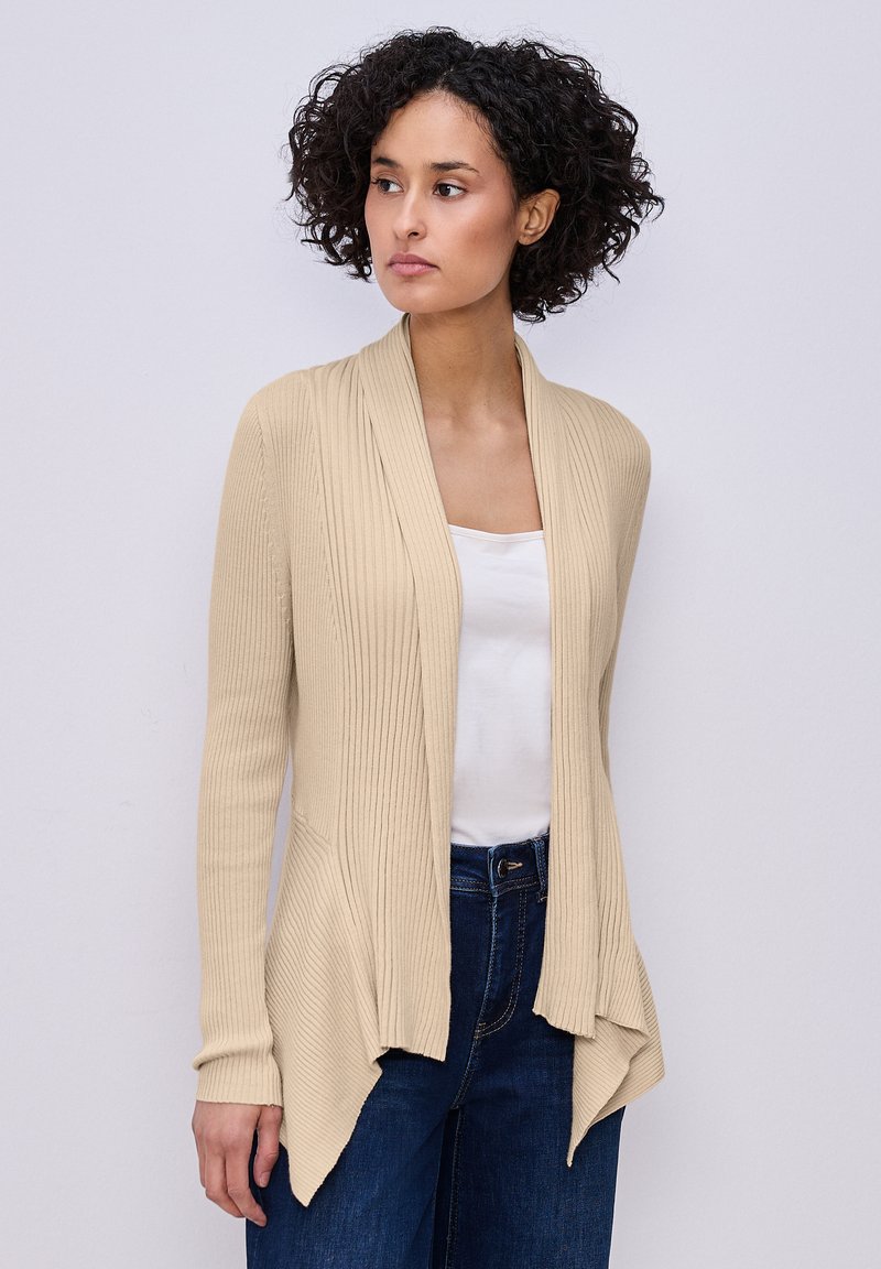 Femme aux cheveux courts et bouclés portant un cardigan beige à côtes ouvert sur le devant, un haut blanc et un jean bleu foncé, debout devant un fond clair uni.