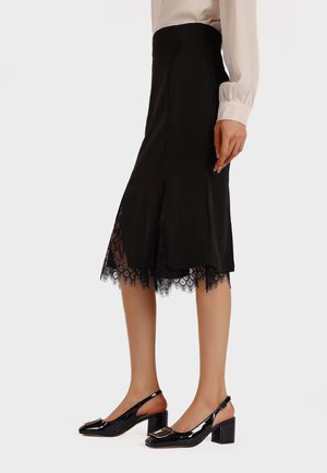 Jupe noire avec un ourlet en dentelle, longueur mi-mollet, fentes sur les côtés et texture lisse. Associée à des escarpins à talons en vernis noir.