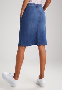 Falda de mezclilla en azul medio con corte recto, hasta la rodilla, con pasadores para cinturón y bolsillos laterales. Combinada con zapatillas de plataforma blancas.