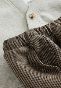 Grijze stoffen top met een knoopdetail, gecombineerd met bruin geruite elastische taillebroek, die zowel gladde als gestructureerde materialen laat zien.