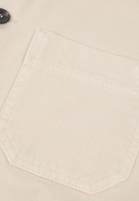 Beige cotton fabric with a diagonal ribbed texture. Features a left chest pocket with triangular stitching and a small logo detail.