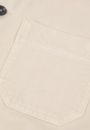 Beige cotton fabric with a diagonal ribbed texture. Features a left chest pocket with triangular stitching and a small logo detail.