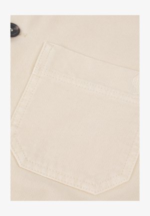 Tessuto di cotone beige con una trama a costina diagonale. Presenta una tasca sul petto sinistro con cuciture a triangolo e un piccolo dettaglio del logo.