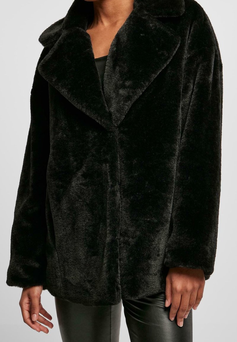 Personne portant un manteau en fausse fourrure noir pelucheux avec un large col et des manches longues, associé à un pantalon en cuir noir.