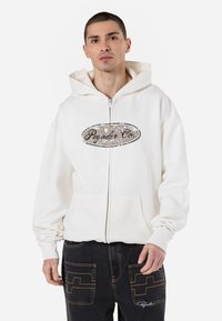 White zip-up hoodie with a front pocket and a graphic logo on the chest. Features a layered hood and ribbed cuffs. Worn with dark jeans.