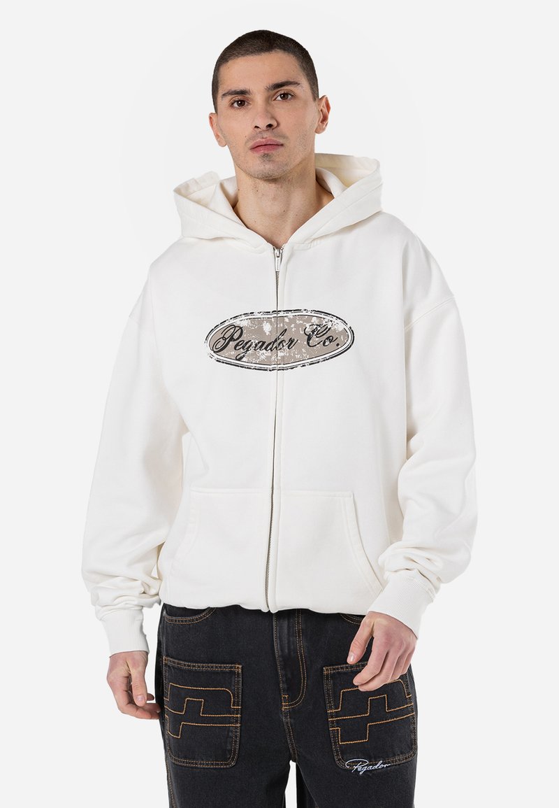 White zip-up hoodie with a front pocket and a graphic logo on the chest. Features a layered hood and ribbed cuffs. Worn with dark jeans.