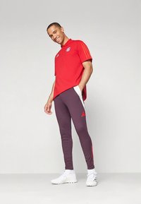 Piros, rövid ujjú galléros póló, Adidas logóval, sötétkék melegítőnadrággal, piros oldalsávokkal és fehér részletekkel. Fehér sportcipő.