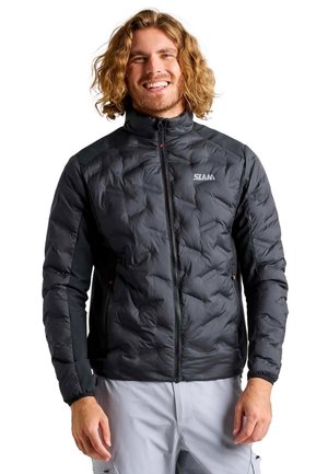 Un hombre sonriente con el pelo ondulado hasta los hombros luce una chaqueta negra acolchada con cremallera y unos pantalones gris claro sobre un fondo blanco.