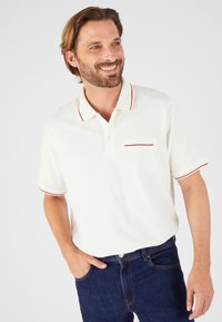 Camisa polo blanca con un acento rojo en el cuello y en el bolsillo. Mangas cortas, hecha de un tejido suave. Llevada con unos vaqueros oscuros.