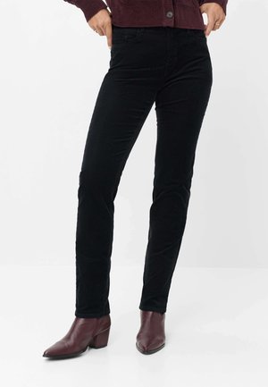 Person iført sorte slim-fit bukser med burgunderrøde ankelstøvler med hæl og en burgunderrød sweater, stående med hænderne i lommerne.