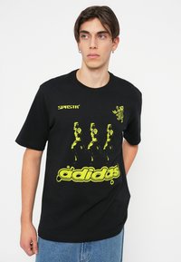 Melna kokvilnas T-krekls ar dzeltenām grafikām, tostarp tekstu "SPRST", skrienošu figūru attēliem un "adidas" logotipu ar pilēšanas efektu.
