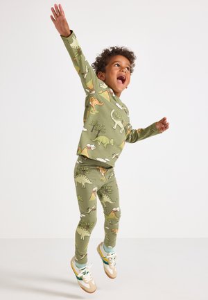Kind dat springt met opgestoken armen, gekleed in groene pyjama's bedrukt met dinosaurussen, vulkanen en bomen, en witte sneakers.
