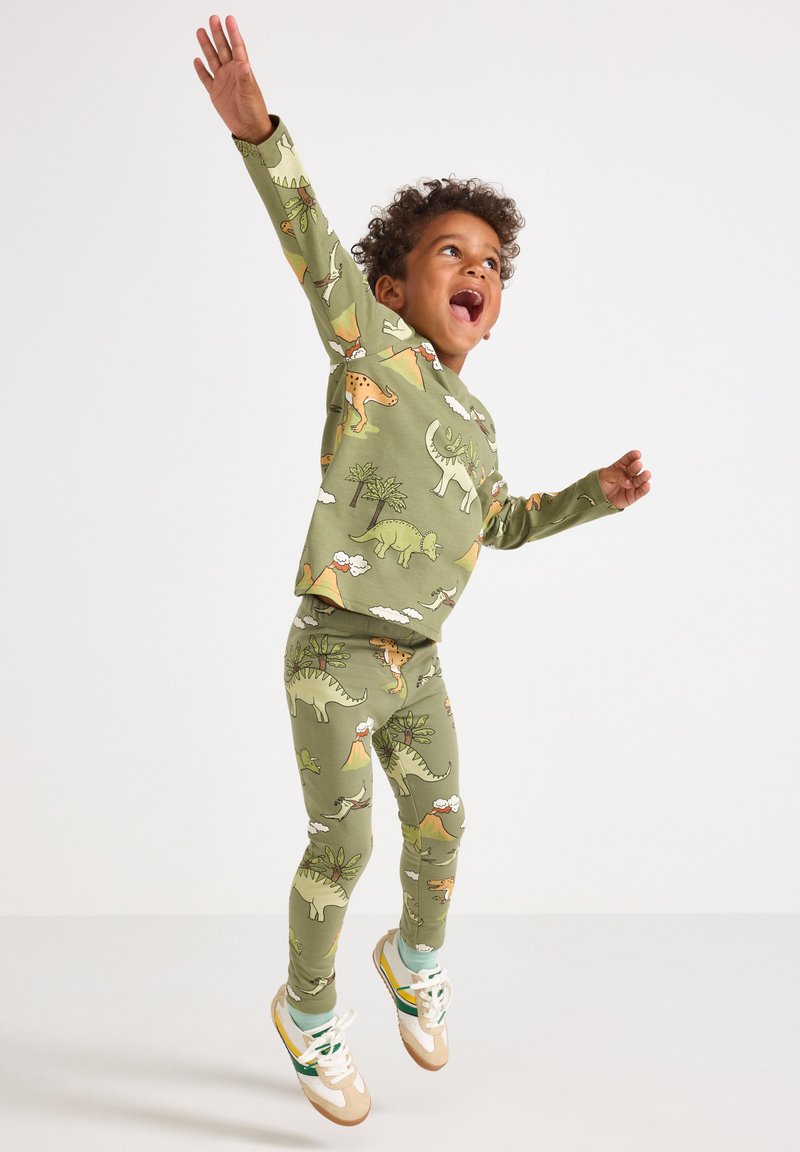 Bambino che salta con le braccia alzate, indossando un pigiama verde stampato con dinosauri, vulcani e alberi, e sneakers bianche.