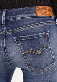 Jeans de mezclilla en azul oscuro con un acabado desgastado. Presenta un patrón decorativo de costura en el bolsillo trasero y una etiqueta de cuero marrón en la cintura.