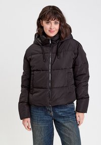 Veste matelassée noire avec capuche, fermeture éclair à l'avant et détails à cordon de serrage. Présente une coupe carrée et une texture matelassée et rembourrée. Associée à un jean bleu.