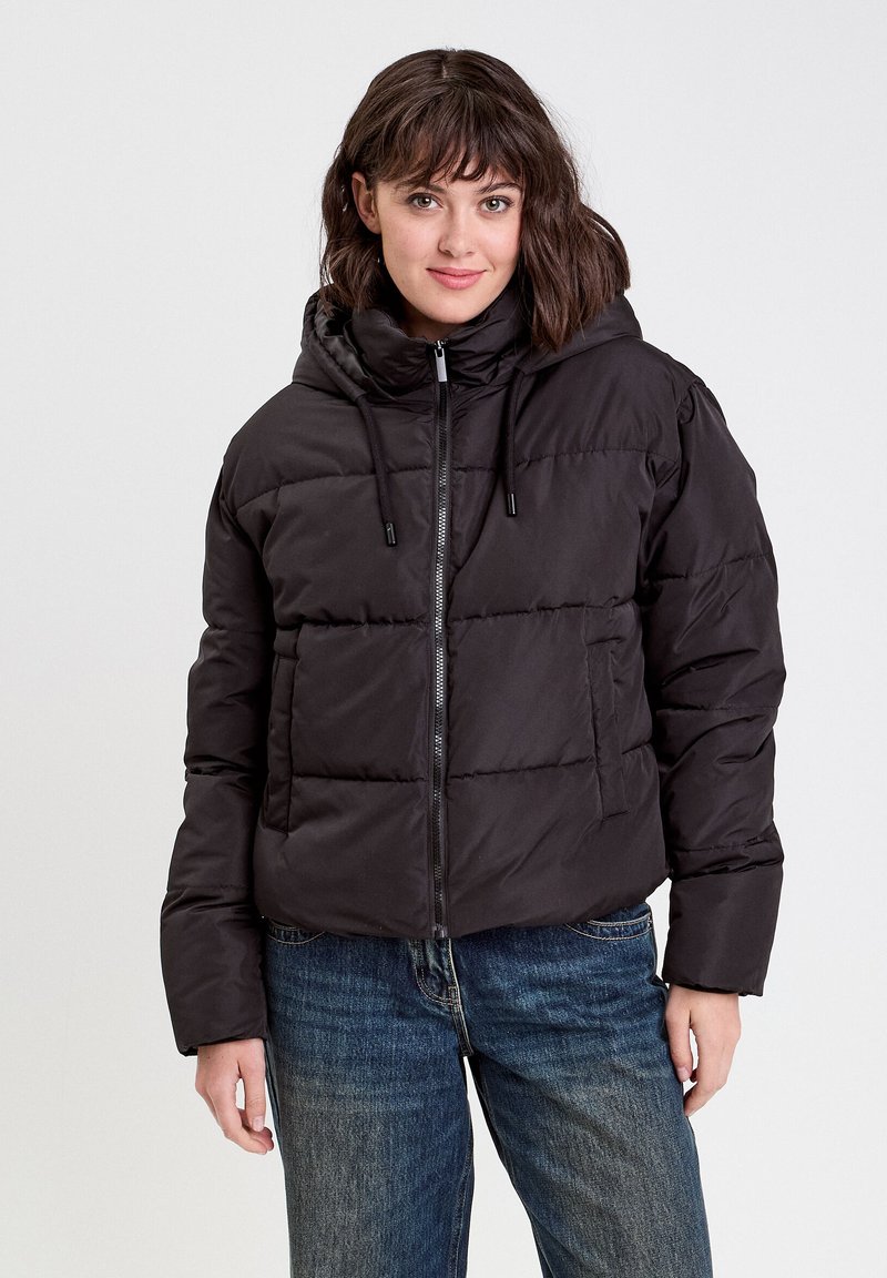 Veste matelassée noire avec capuche, fermeture éclair à l'avant et détails à cordon de serrage. Présente une coupe carrée et une texture matelassée et rembourrée. Associée à un jean bleu.