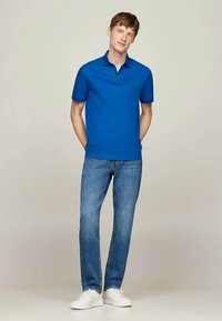 Ung mann står med hendene bak ryggen, iført en blå polo-skjorte, blå jeans og hvite joggesko på en ensfarget bakgrunn.