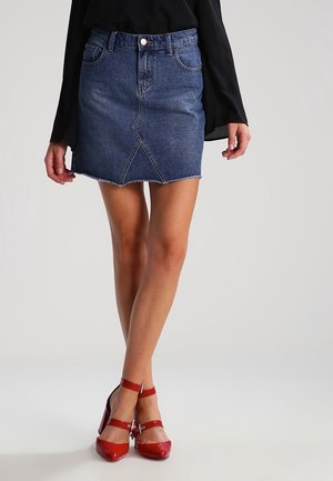 Kvinde iført en blå denim mininederdel med detalje i frontsøm, sort langærmet top og røde højhælede sko med ankelrem.