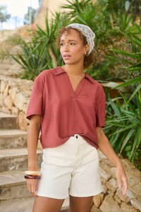 Jeune femme portant un haut à manches courtes couleur rouille, un short blanc, un foulard en dentelle blanche sur la tête et des bracelets, debout près d'escaliers en pierre et de plantes vertes.
