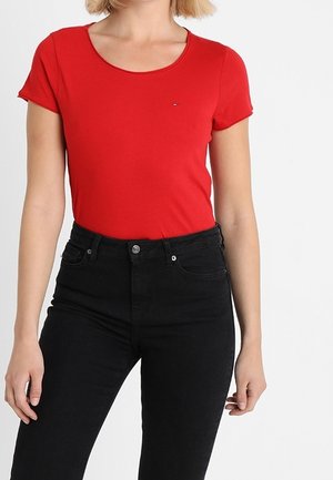 Mujer con una camiseta ajustada de manga corta roja y vaqueros negros de cintura alta, de pie frente a un fondo blanco liso.