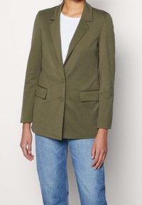 Blazer vert olive avec un col tailleur, deux boutons et des poches latérales, fabriqué dans un tissu doux et léger ; associé à un haut blanc et un jean bleu.