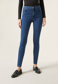 Jeans ajustados de tiro alto en denim azul oscuro con cinco bolsillos, cierre de botón y un ligero desvanecimiento en la parte frontal. Combinados con mocasines negros.