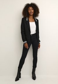 Blazer noir ajusté avec des accents rayés blancs, porté par-dessus un top blanc. Associé à un jean skinny noir et des bottines noires pointues.