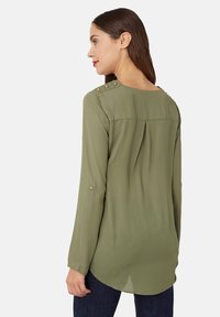 Mujer con cabello castaño oscuro que lleva una blusa de manga larga verde oliva con botones dorados en los hombros y las mangas, combinada con jeans oscuros.