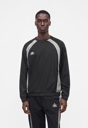 Άνδρας που στέκεται φορώντας μαύρη φόρμα Umbro με γκρι πλαϊνές ρίγες και ελαστικά τελειώματα στα μανίκια και στα πόδια, πάνω σε λευκό απλό φόντο.