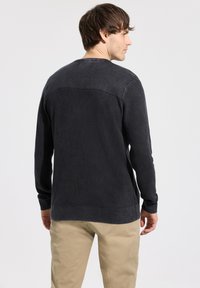 Pull gris foncé à manches longues avec un design côtelé texturé, un panneau d'épaule lisse contrastant et des poignets ajustés, associé à un pantalon beige.