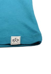 Teal bomulds T-shirt med foldet kant, prydet med et cremefarvet mærke med sort tekst og krydsede linjer. Blød tekstur, enkelt design.