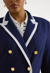 Artimas asmuo, dėvintis tamsiai mėlyną švarką su baltomis apvadomis, auksiniais mygtukais, baltą polo marškinėlį su tamsiai mėlynomis juostelėmis ir auksinius kabančius auskarus.