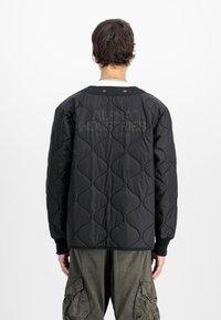 Chaqueta negra acolchada con un patrón de costura curvo, puños acanalados y un logo en la parte posterior. Llevada sobre pantalones cargo verde oliva.