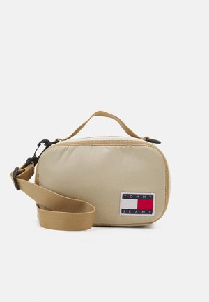 Light beige fabric bag with a zip closure, carrying handle, and adjustable strap. Features a prominent "TOMMY JEANS" logo patch.