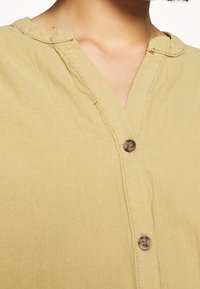 Camicia khaki con bottoni, scollatura a V, tessuto testurizzato e dettagli dei bottoni visibili sul davanti. Leggeri pizzichi accentuano il suo stile casual.