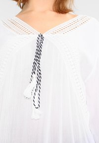 Blusa bianca con scollatura a V e dettagli in pizzo intricati. Coulisse regolabile in bianco e nero con frange nelcollo.