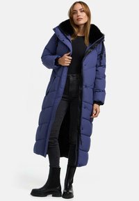 Cappotto lungo blu imbottito con colletto in pelliccia sintetica nera, texture trapuntata, apertura frontale e tasche anteriori. Abbinato a jeans neri e stivaletti neri.