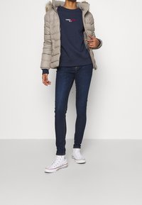 En pufferjacka i ljusgrått med huva med pälsdetalj, en marinblå sweatshirt med logotyp, mörka skinny jeans och vita högskor.