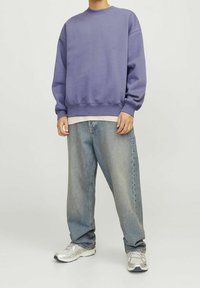 Person trägt ein locker sitzendes lila Sweatshirt, ein hellbeiges Unterhemd, weite, ausgebleichte blaue Jeans und silber-weiße Turnschuhe und steht vor einem schlichten Hintergrund.