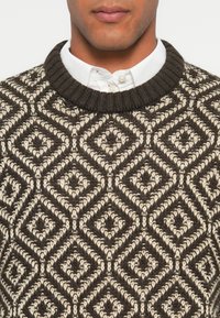 Vīrietis valkā tumši pelēku un baltu strukturētu adītu džemperi ar dimanta rakstu, kas uzvilkts virs baltas pogas krekla.