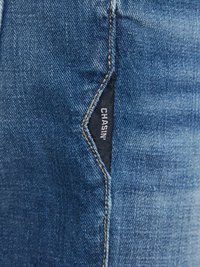 Tecido de denim azul com uma textura sutil, apresentando costuras visíveis e uma etiqueta com "CHASIN'" posicionada na costura lateral.