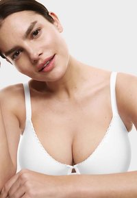 Bílá podprsenka s nastavitelnými ramínky, měkkou tkaninou, hladkou texturou a klíčovým otvorem uprostřed. Kalíšky mají vlnité okraje.