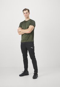 Olive zaļa sporta T-krekls ar melnajām sānu svītrām, apvienots ar melniem sporta biksēm, kuru priekšpusē ir pelēks logo, papildināts ar melnām ikdienas apavām.