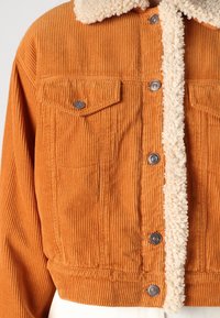 Giacca corta in velluto a coste color arancione bruciato con collo soffice color avorio, dotata di due tasche sul petto e bottoni a pressione argentati.